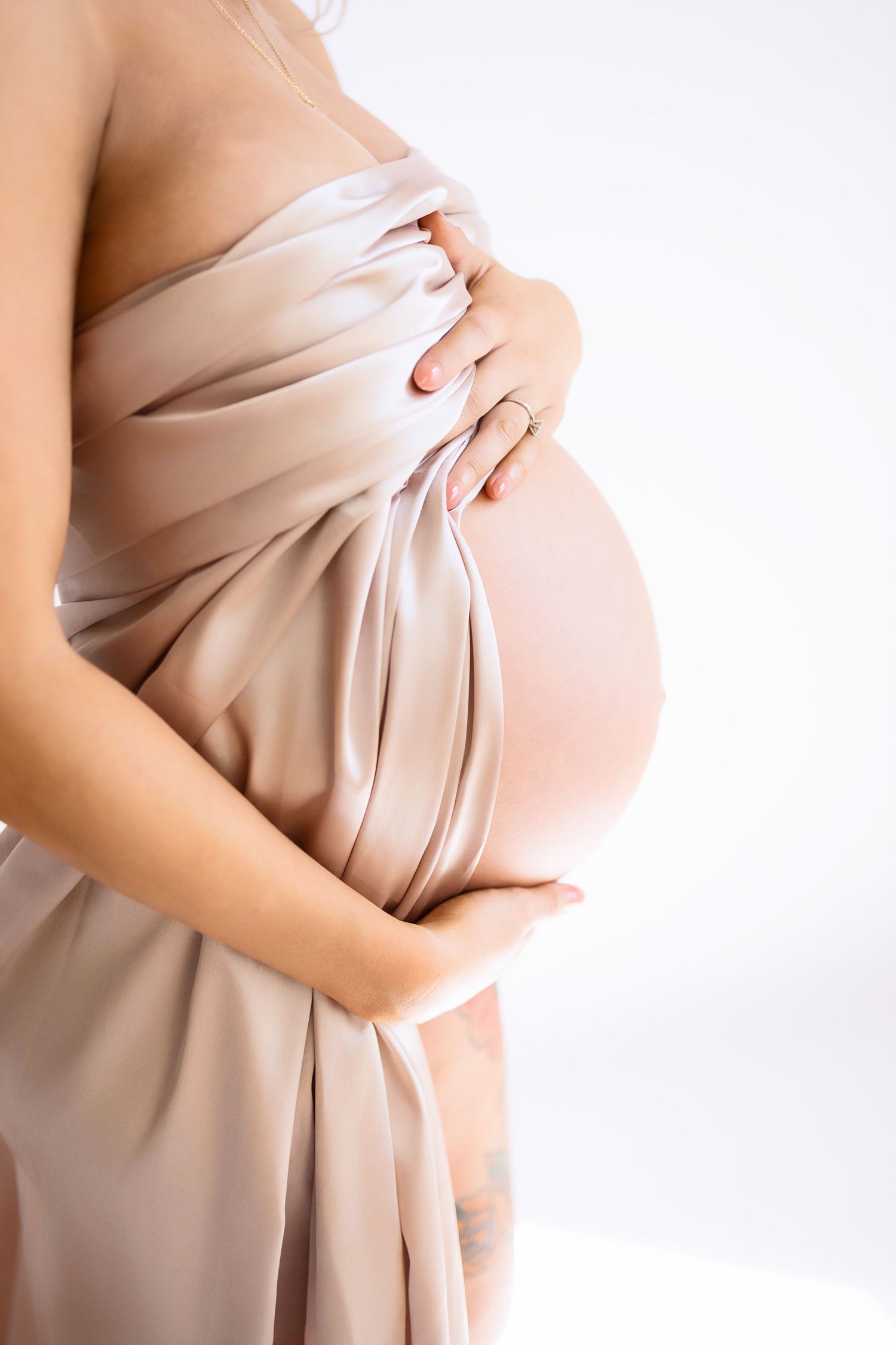 A pregnant woman draped in soft beige fabric gently holds her belly against a white background.