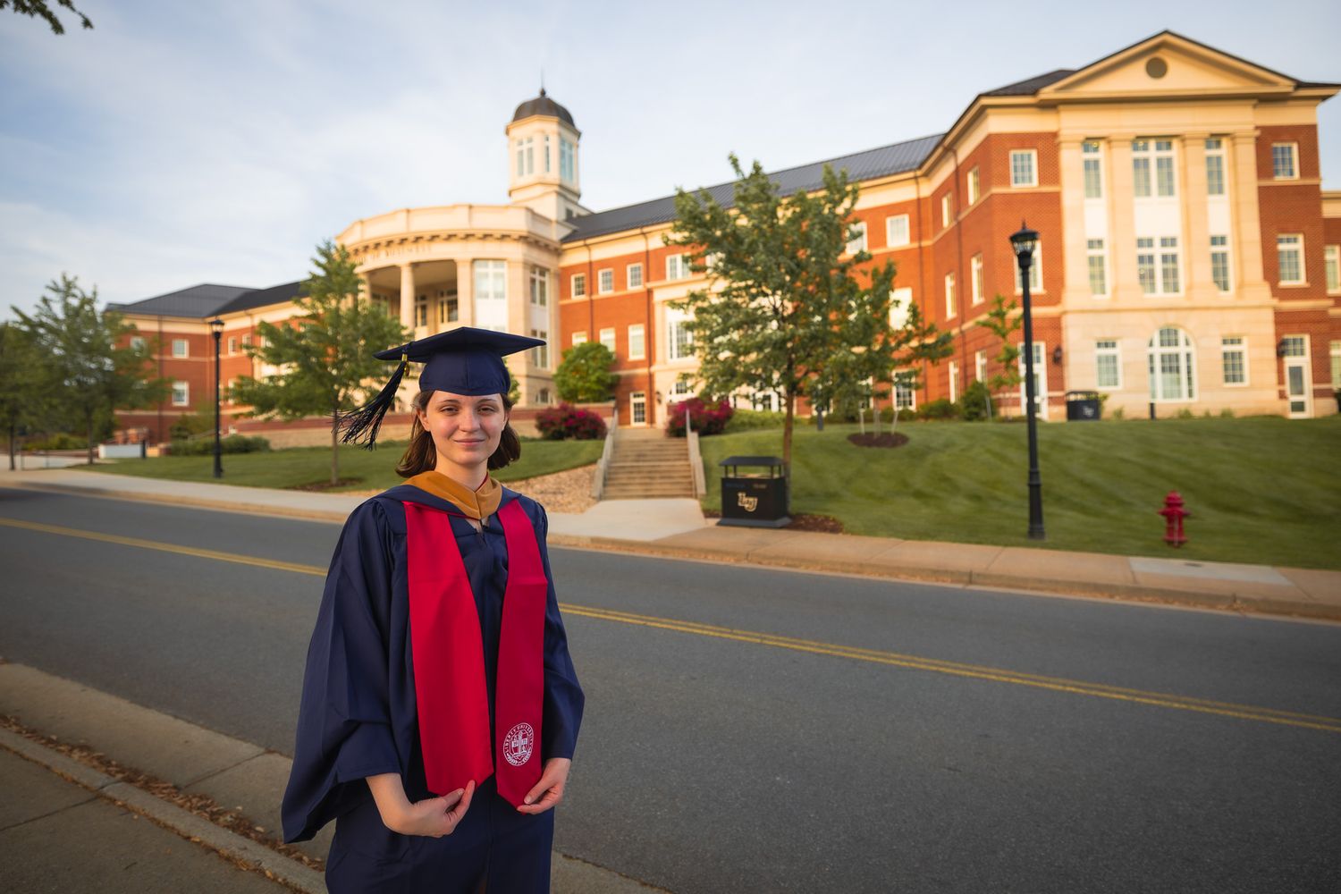 Graduate in cap and gown stands outside brick university buildings on graduation day.