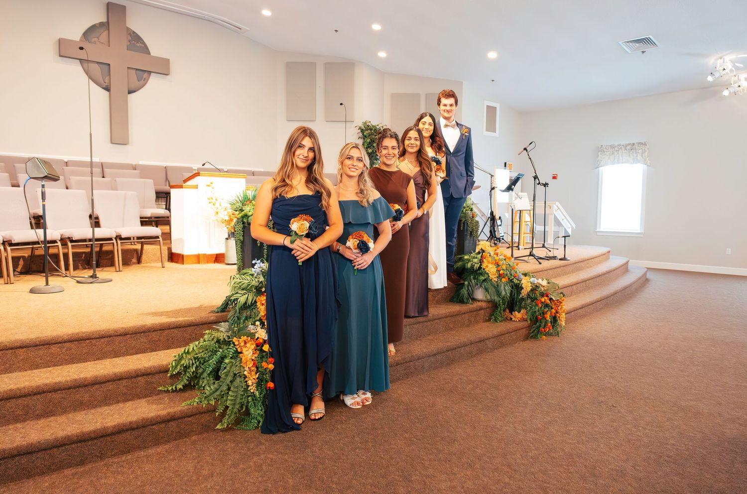 Wedding party in navy blue dresses stands on church altar steps decorated with greenery and floral arrangements.