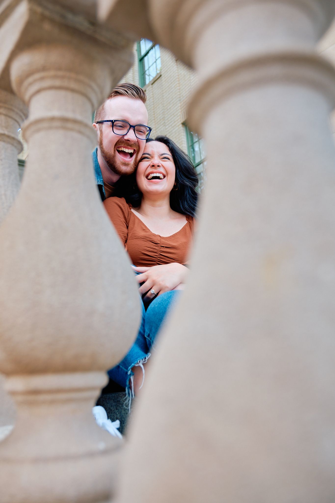 A couple shares joyful laughter while posing between architectural columns in a creative engagement photo series.