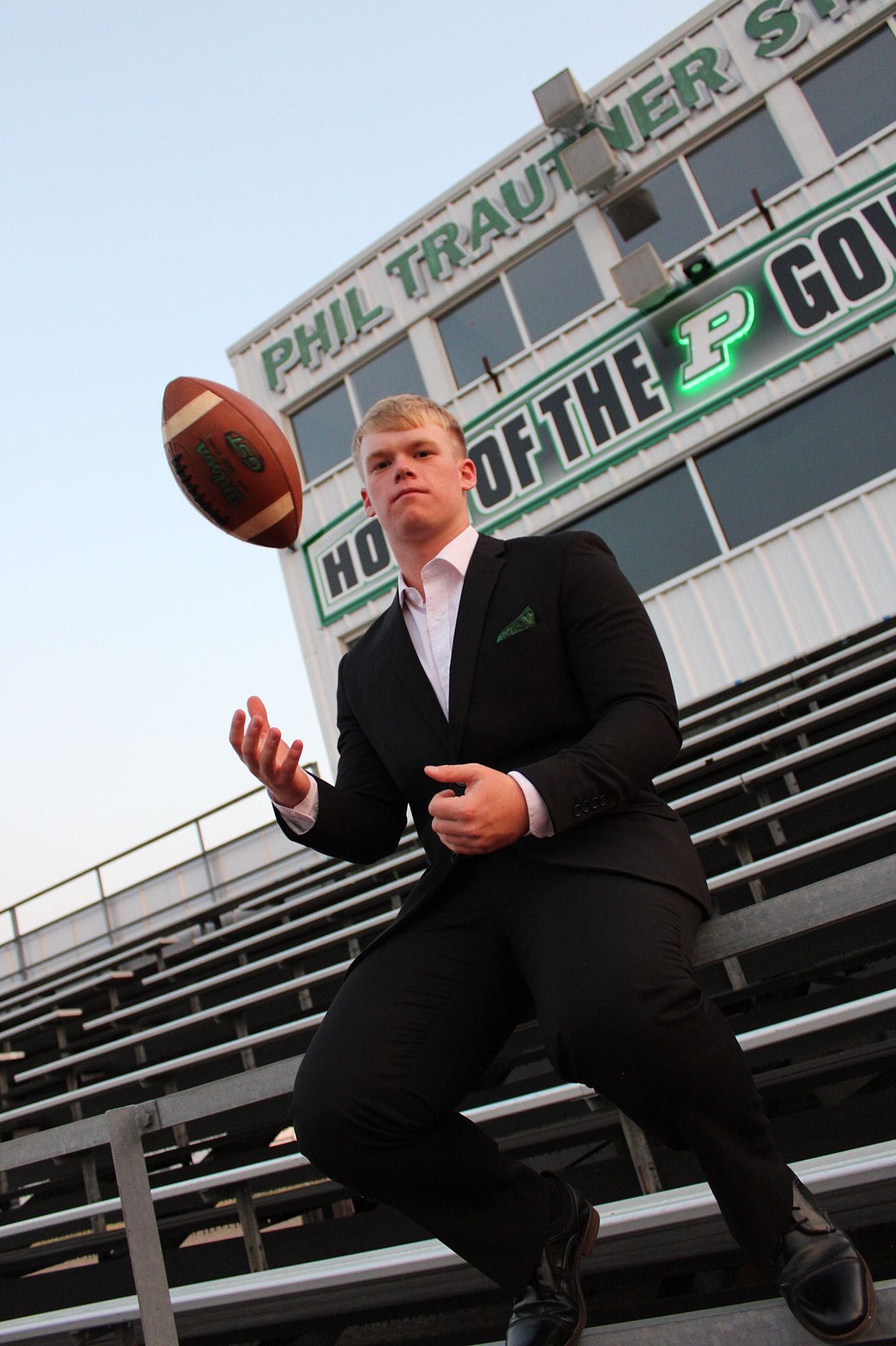 A person in a black suit tosses a football while standing in stadium bleachers with signage visible in the background.