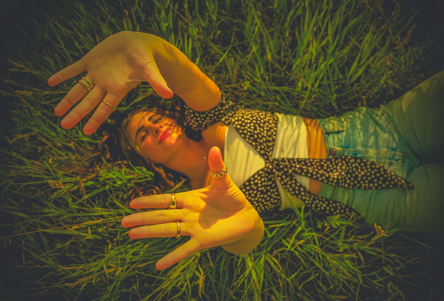 A person lies in tall grass with outstretched hands while wearing a patterned outfit in warm golden lighting.