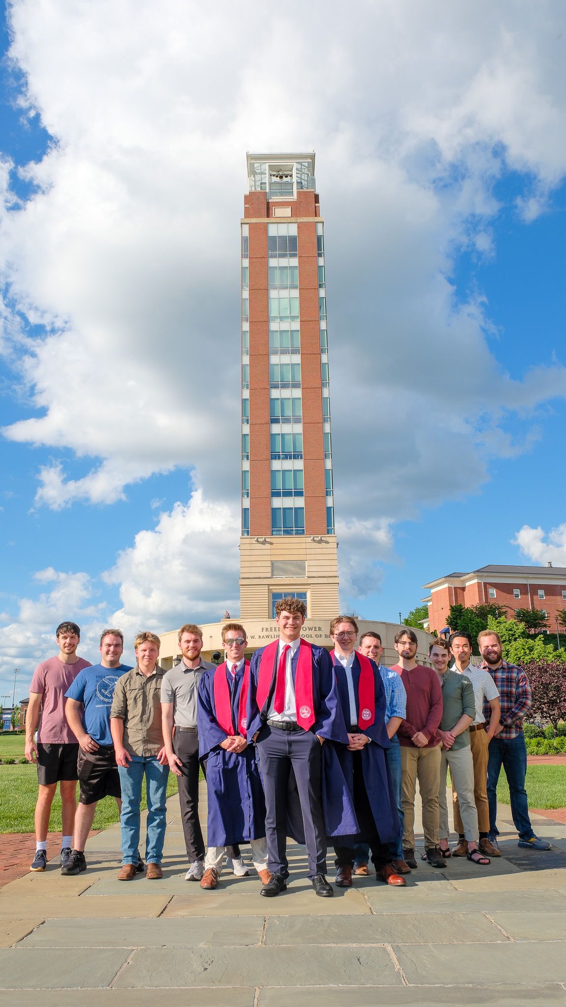 A group stands together in front of a tall brick clock tower on a university campus against a blue cloudy sky.