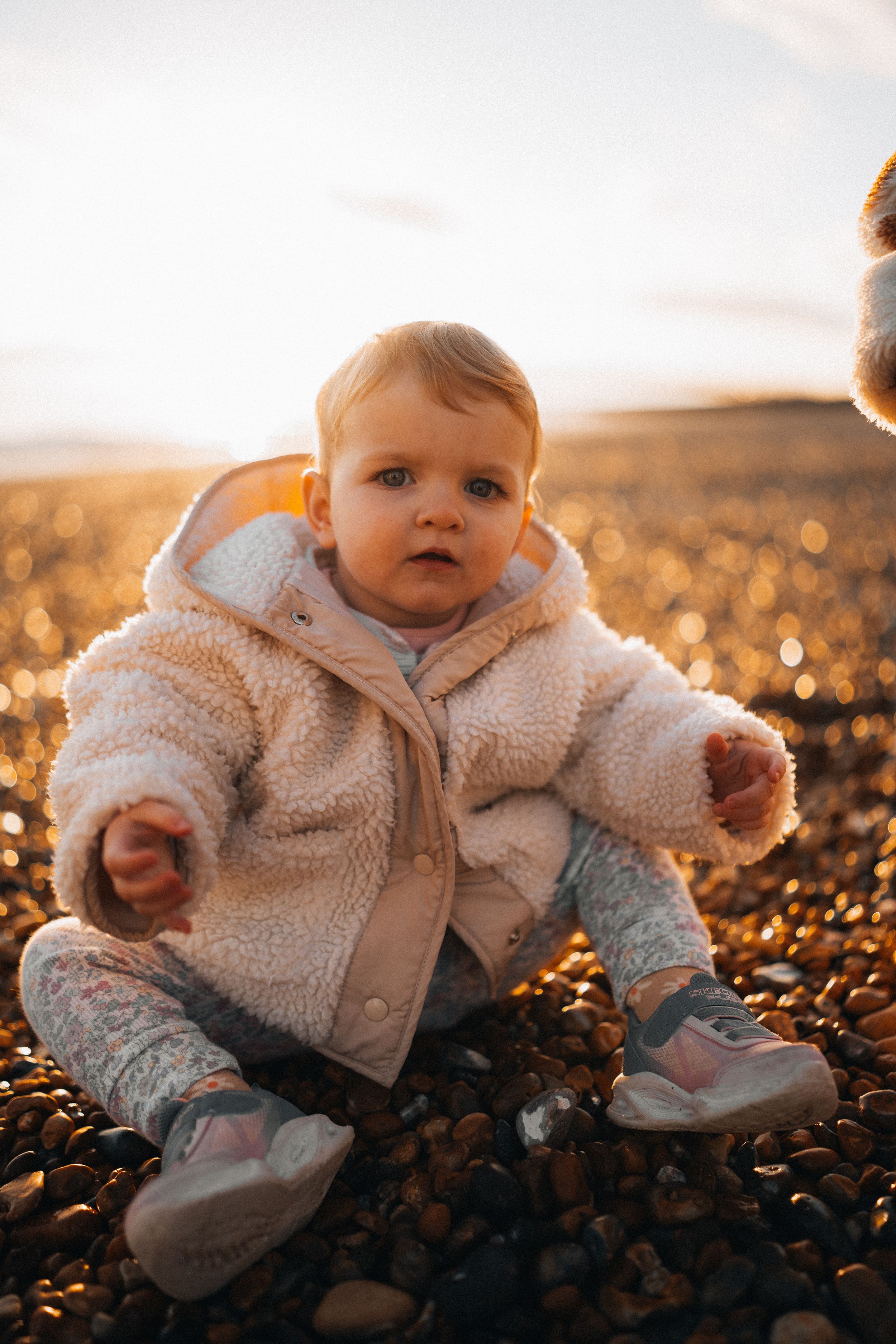 Baby in white fleece outfit sitting on autumn ground at sunset.