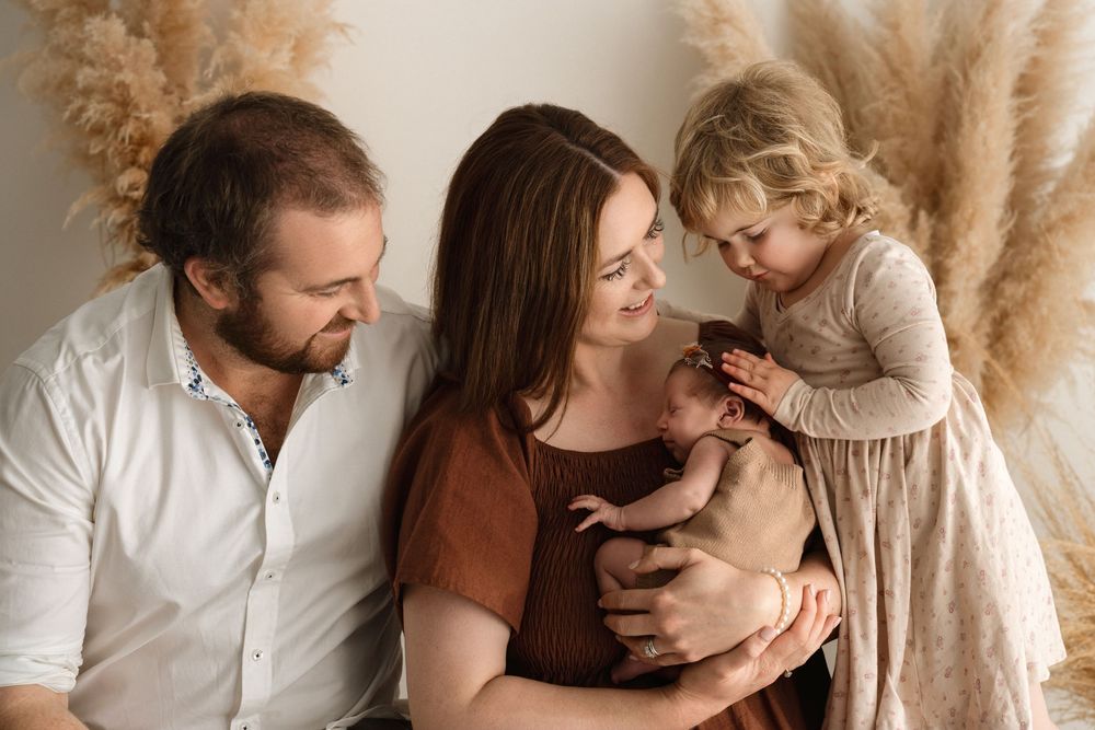 Family of four with parents holding newborn baby while toddler girl leans in, surrounded by pampas grass decor.