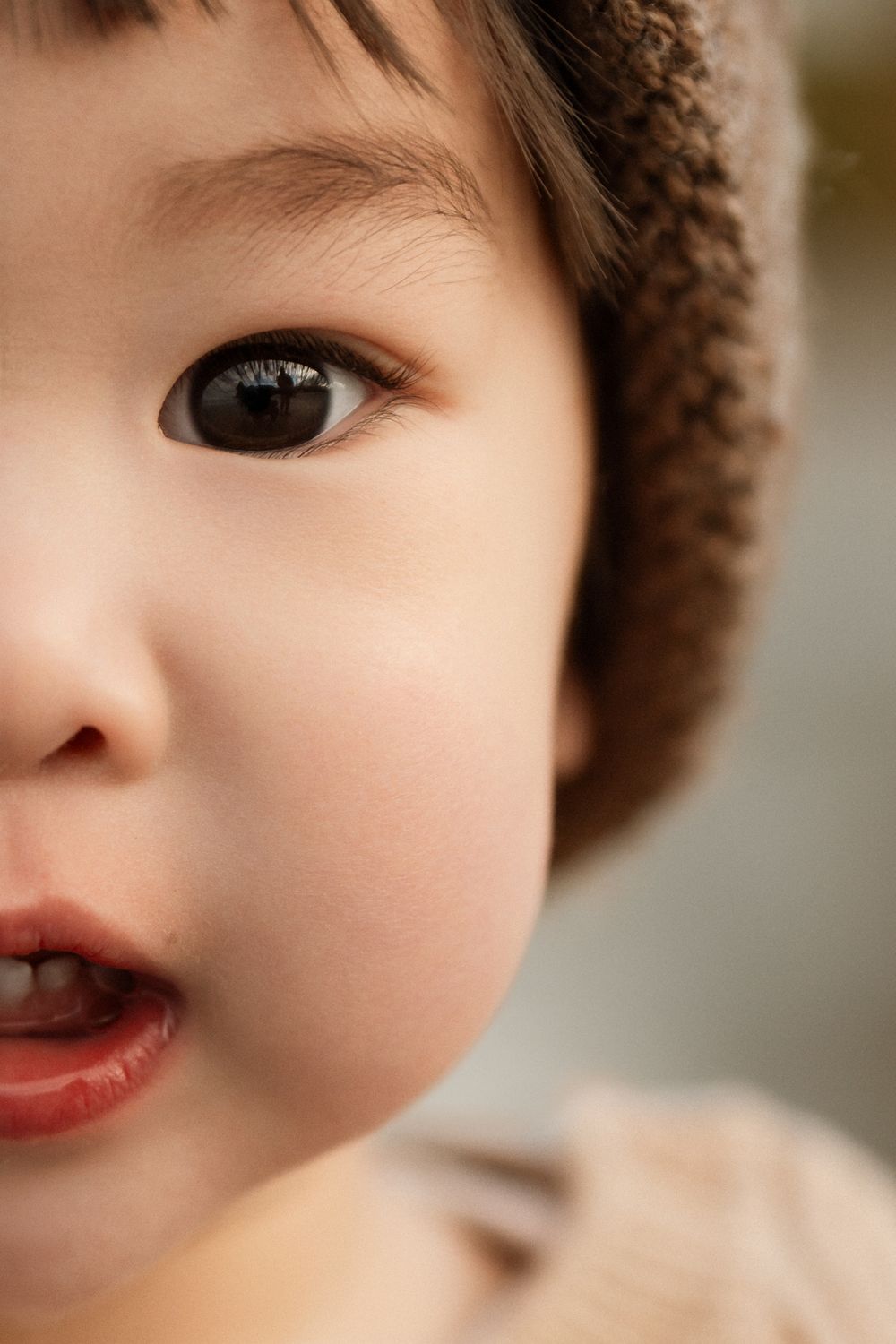 Close-up portrait of a cute Asian toddler with big brown eyes wearing a knit hat, mouth slightly open.