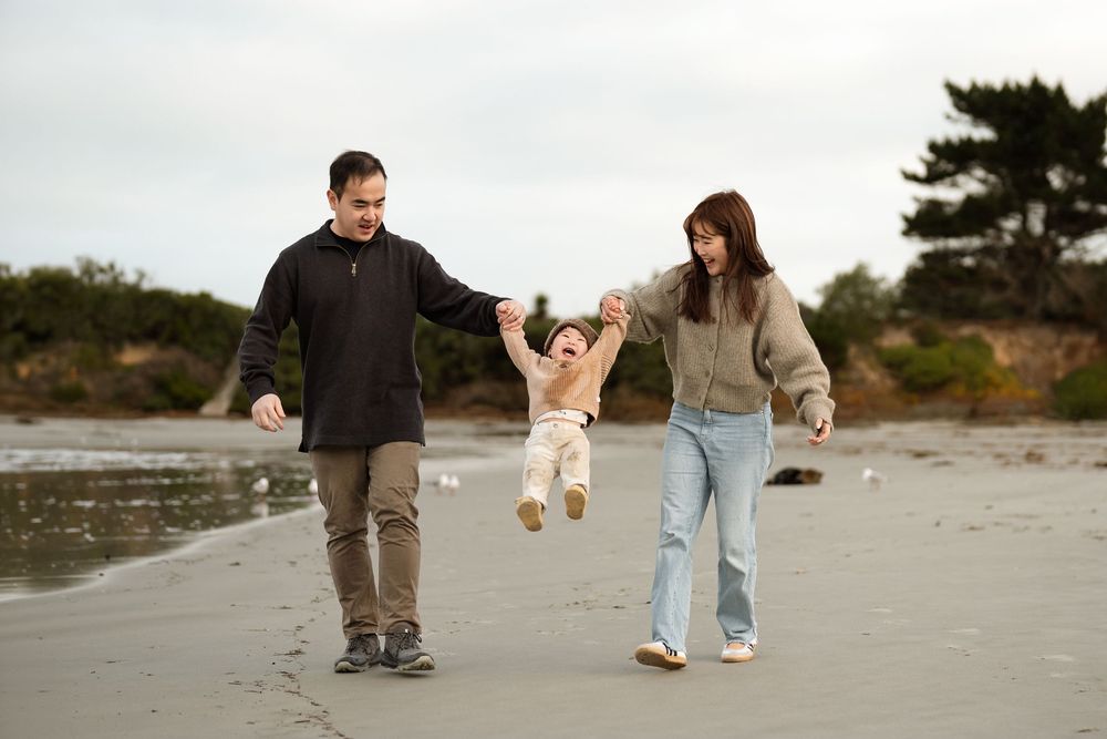 Family swinging baby on beach, parents holding toddler hands, cloudy coastal walk, trees in background.