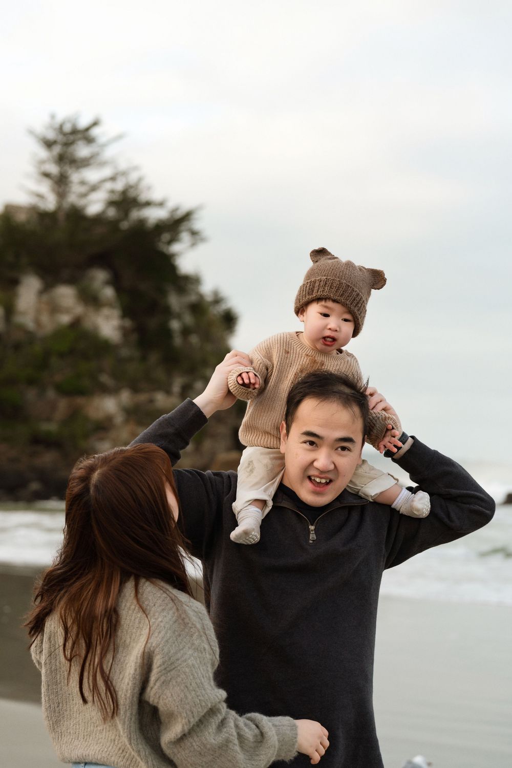 Young Asian family at beach, father carrying baby on shoulders while mother holds child, all smiling outdoors.