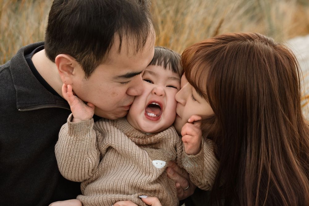 Two parents lovingly kissing their laughing baby on the cheeks outdoors in a heartwarming family portrait.
