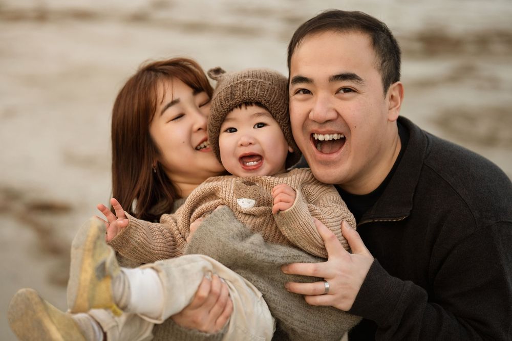 Happy Asian family with laughing baby in knit hat, parents smiling outdoors in a warm, joyful portrait.