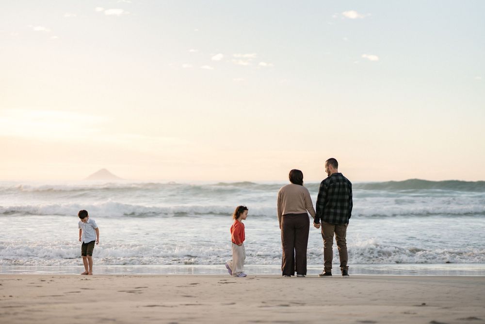 Family of four standing together on a beach at sunset, watching ocean waves with a distant mountain on the horizon.