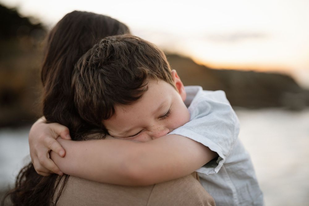 A young boy rests his head on a woman's shoulder in a tender embrace near a beach at sunset.