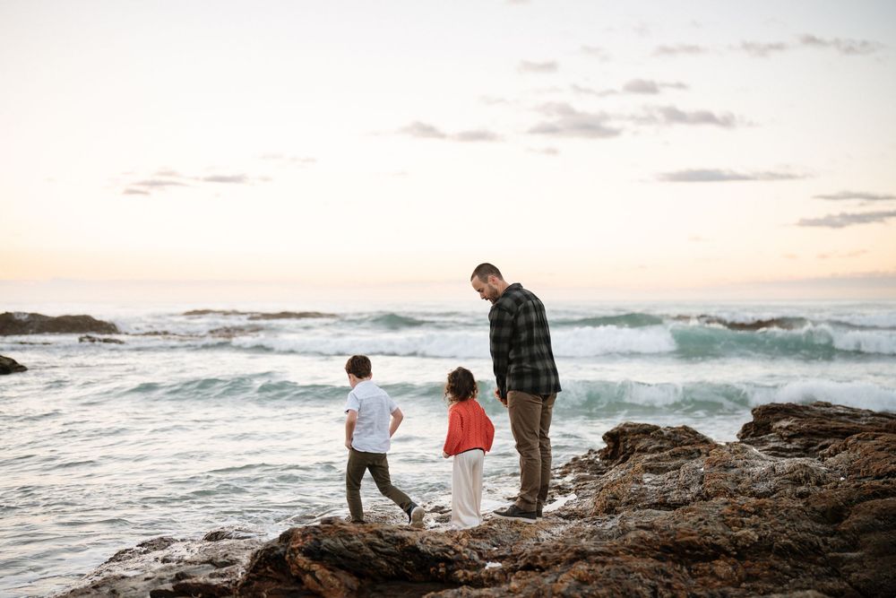 Father and two children standing on rocky shore watching ocean waves at sunset.