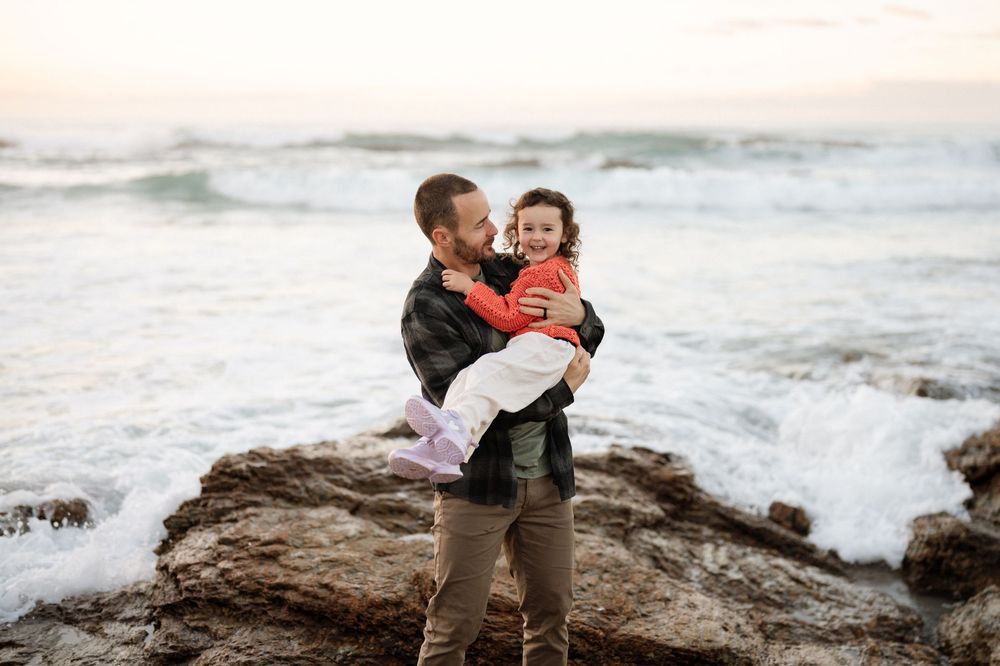 A father joyfully holds his young child on rocky coastal shores with ocean waves crashing behind them at sunset.