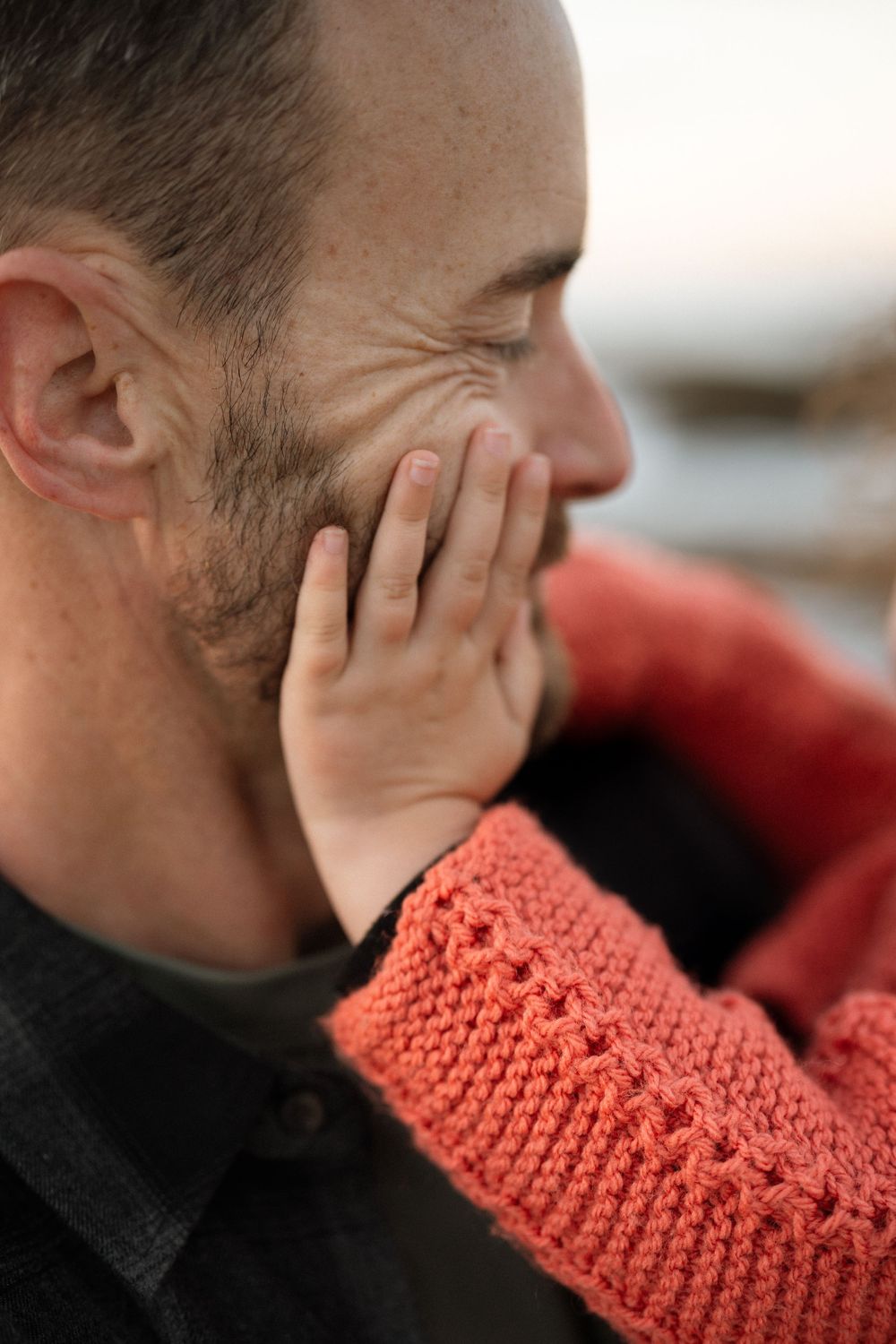 A bearded man in a dark shirt with a small child's hand in a coral knit sleeve touching his face.