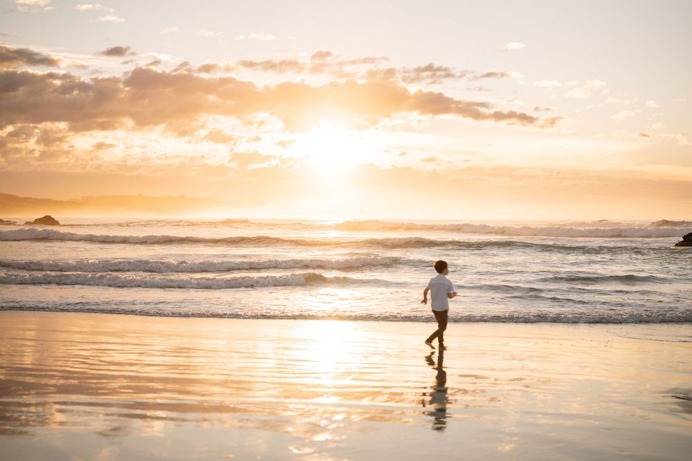 Child running along a golden sandy beach at sunset with glowing orange sky and ocean waves in the background.