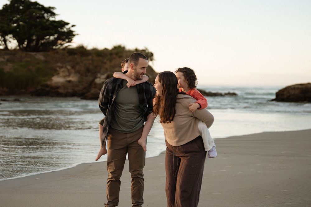 Family of four enjoying a beach walk at sunset, parents carrying children on their backs near the ocean shore.
