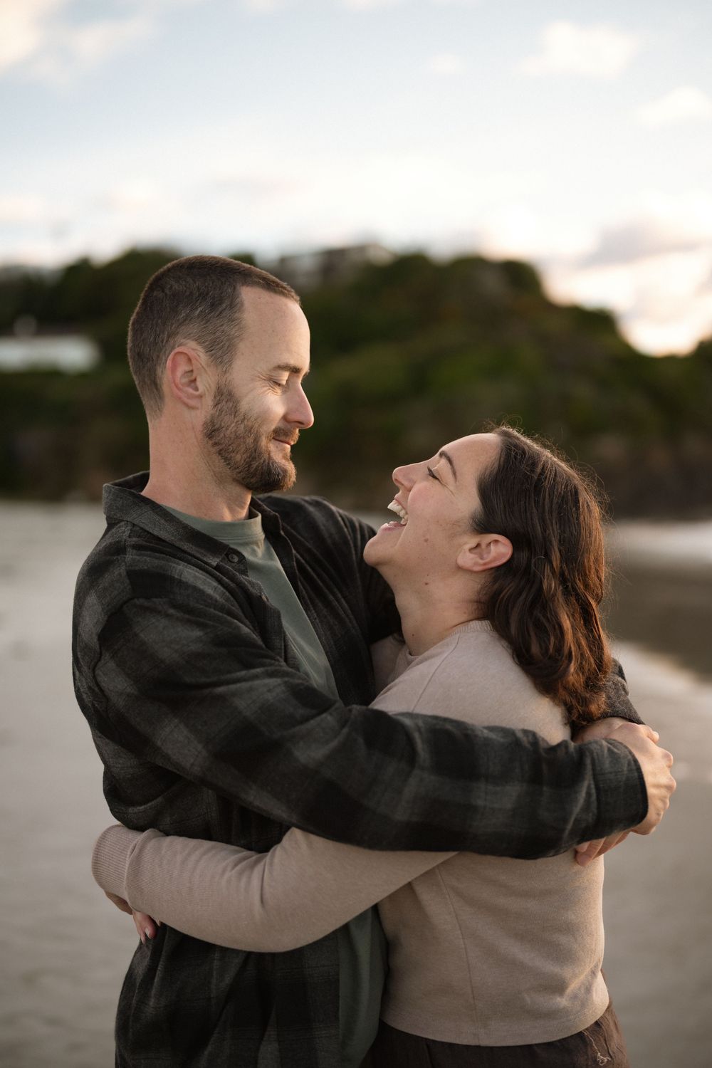 A happy couple embraces and laughs together on a beach at sunset, wearing casual outerwear.