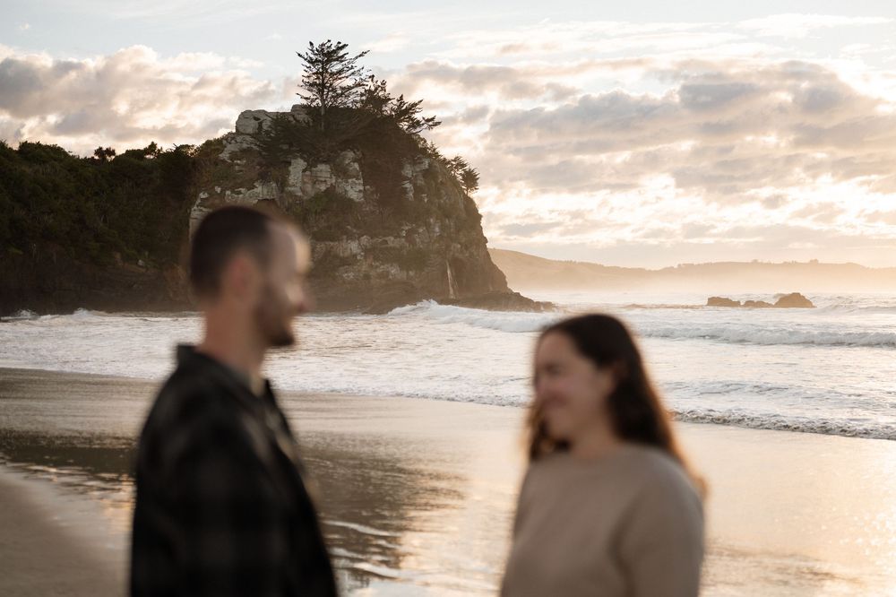 Couple walking on a sandy beach at golden sunset with dramatic rocky headland and ocean waves in background.