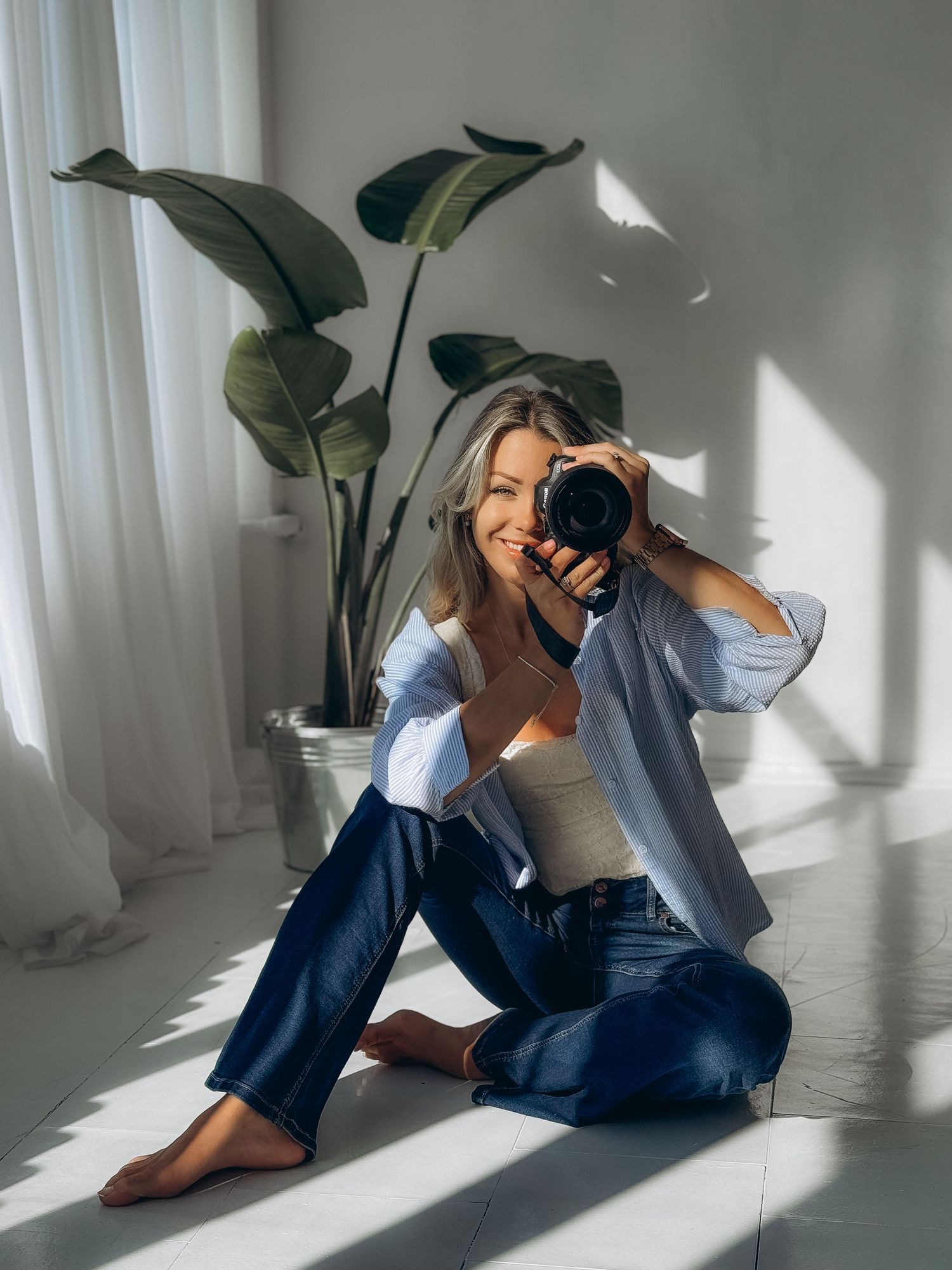 Person in blue jeans and light top sits on floor taking photo with camera near large indoor plant in minimalist setting.