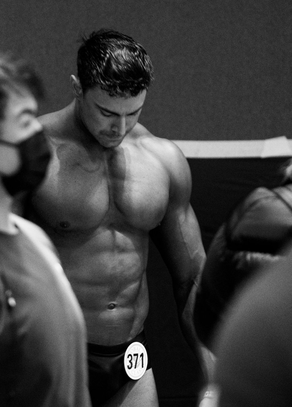 Black and white photo of a muscular male bodybuilder wearing number 371 at a competition.