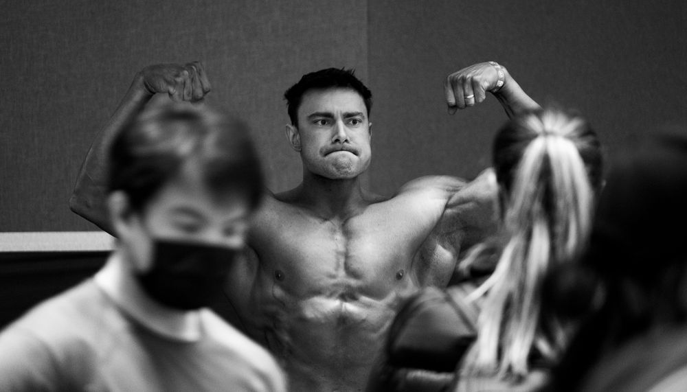 Muscular shirtless man flexing biceps in black and white photo with masked spectators in foreground.