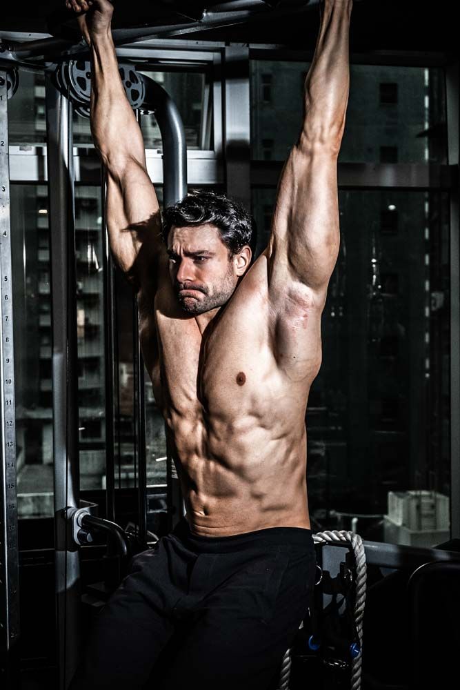 Muscular shirtless man hanging from pull-up bar in gym, displaying defined abs and biceps during intense workout.