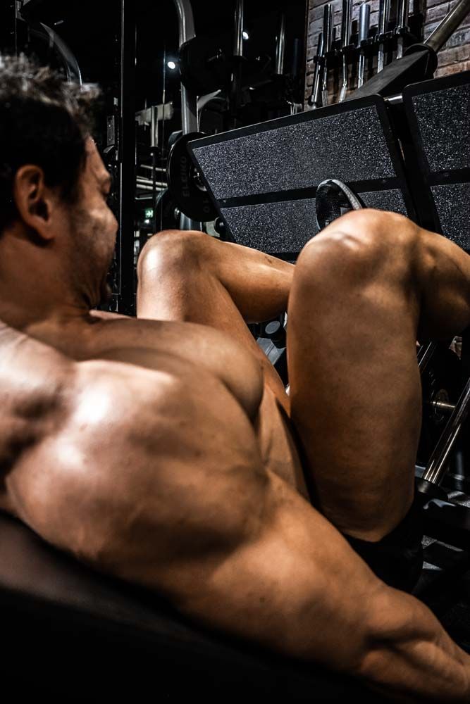 Muscular man performing leg press exercise on gym machine, showcasing defined physique in dramatic dark lighting.