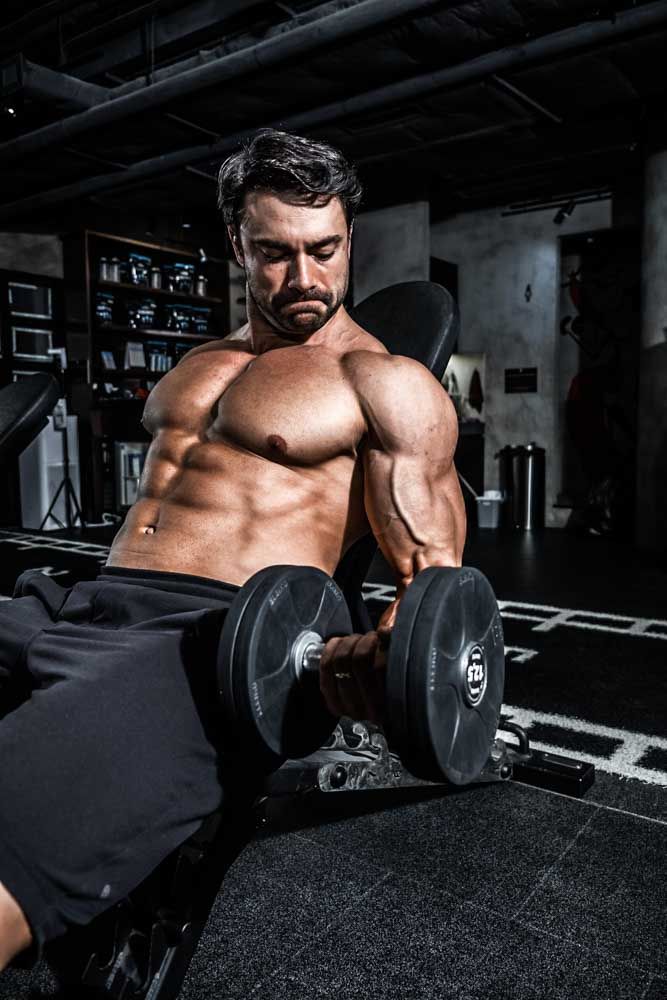 Muscular shirtless man doing dumbbell curls on an incline bench in a dark gym.