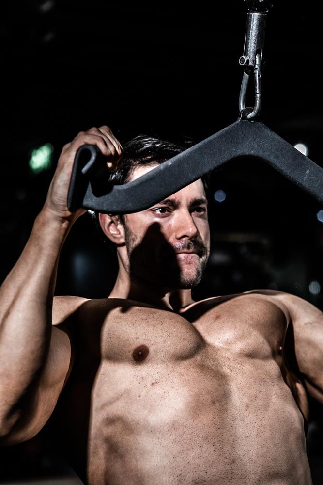 Muscular shirtless man gripping a cable machine attachment in a dark gym, showcasing defined chest and arms.