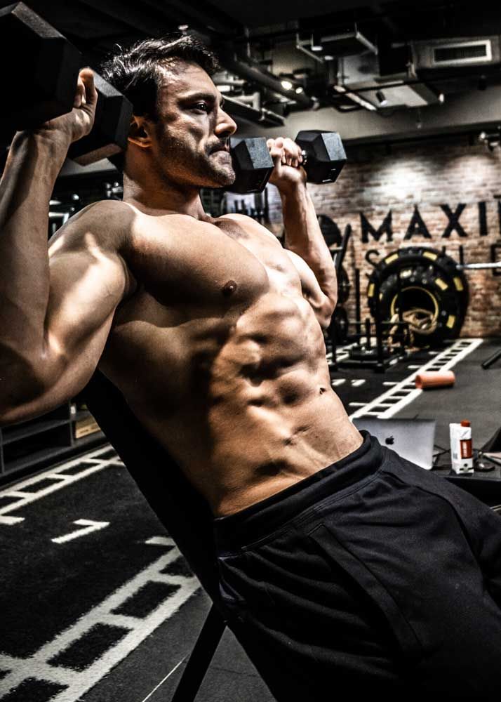Muscular man performing dumbbell exercises on an incline bench in a modern gym, showcasing defined abs and biceps.