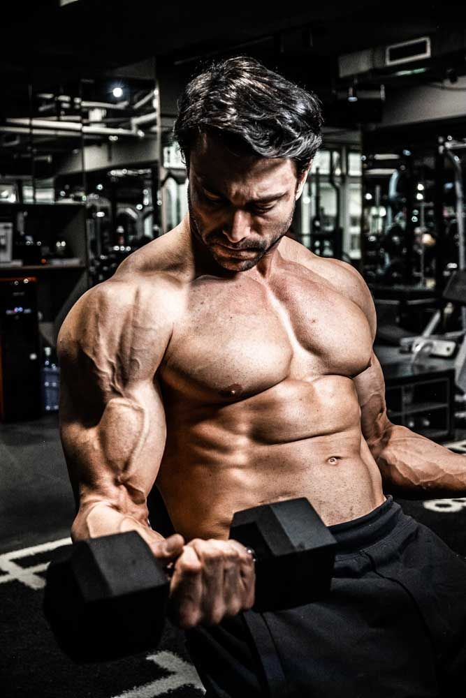 Muscular shirtless man doing dumbbell curls in a dark gym, showcasing defined abs and biceps.