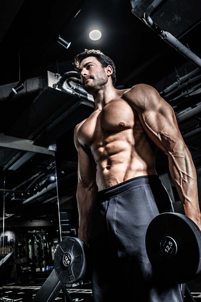 Muscular shirtless man holding dumbbells in a dark gym, showcasing defined abs and arms under dramatic lighting.