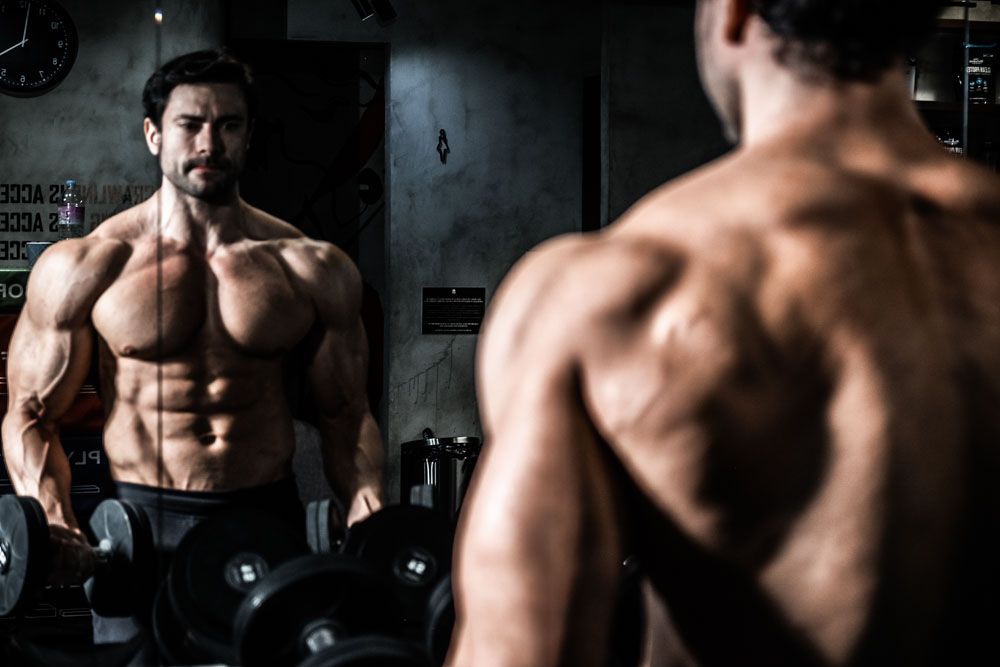 Muscular bodybuilder flexing in gym mirror showing defined back and chest muscles under dramatic lighting.