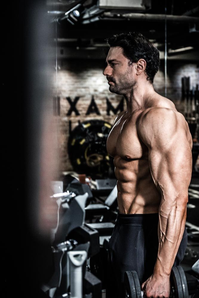 Muscular shirtless man holding dumbbells in a dark gym, showcasing defined abs and arms in a dramatic side profile pose.