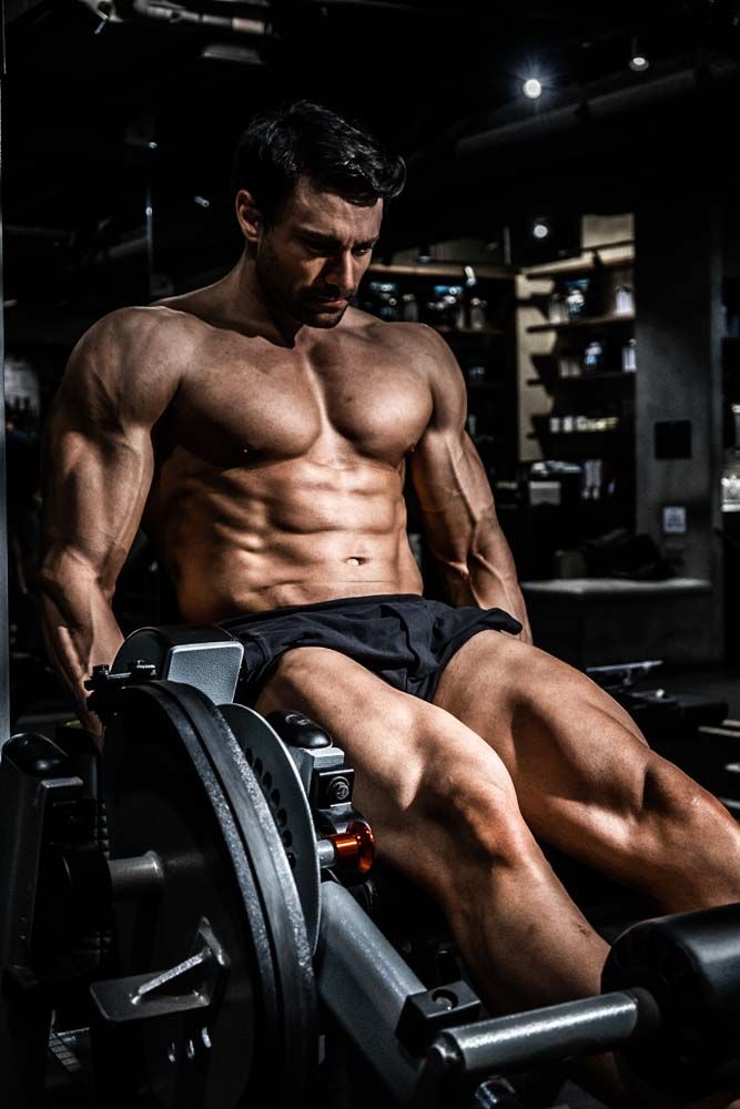 Muscular shirtless man performing leg press exercise on machine in dark gym, showcasing defined physique and strength.