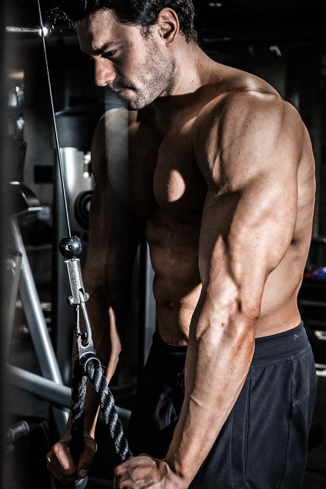Muscular shirtless man performing tricep pulldown exercise on cable machine in dark gym setting.