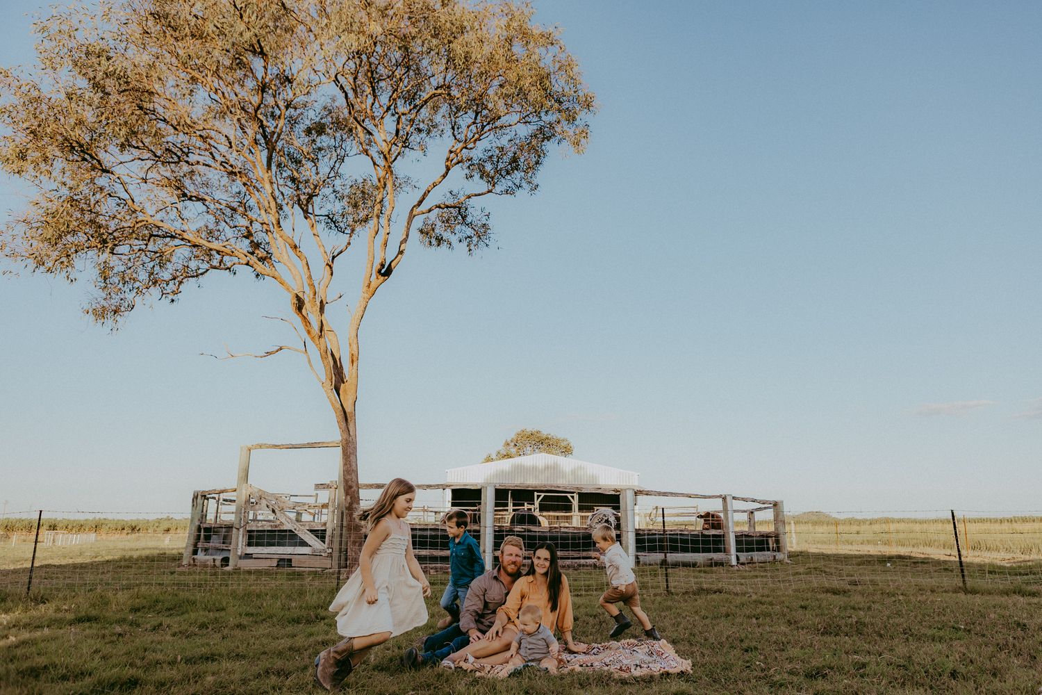 Joyful family posing playfully under a sprawling gum tree on a sunny rural property with a marquee behind.