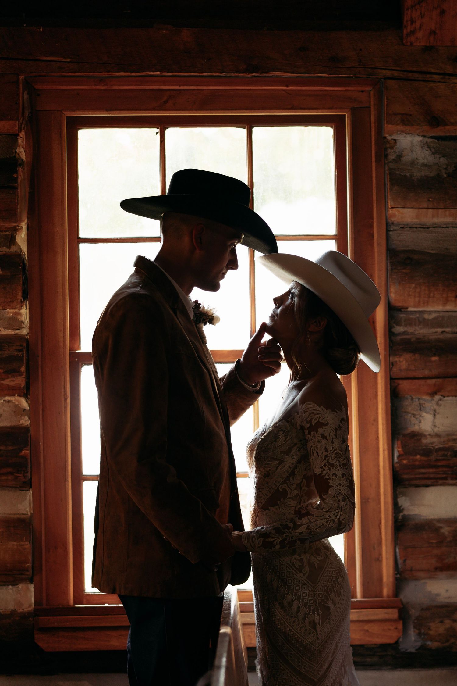 Silhouette of bride and groom in cowboy hats sharing romantic moment by rustic log cabin window.