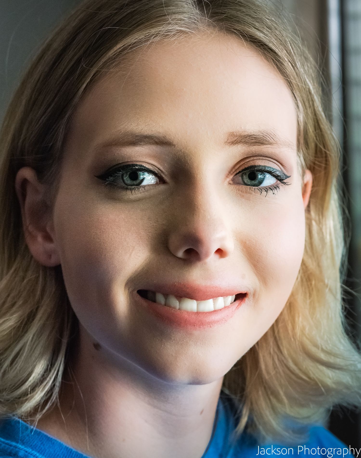 Close-up portrait of a smiling young blonde woman with green eyes and teal eye makeup, photographed by Jackson Photography.