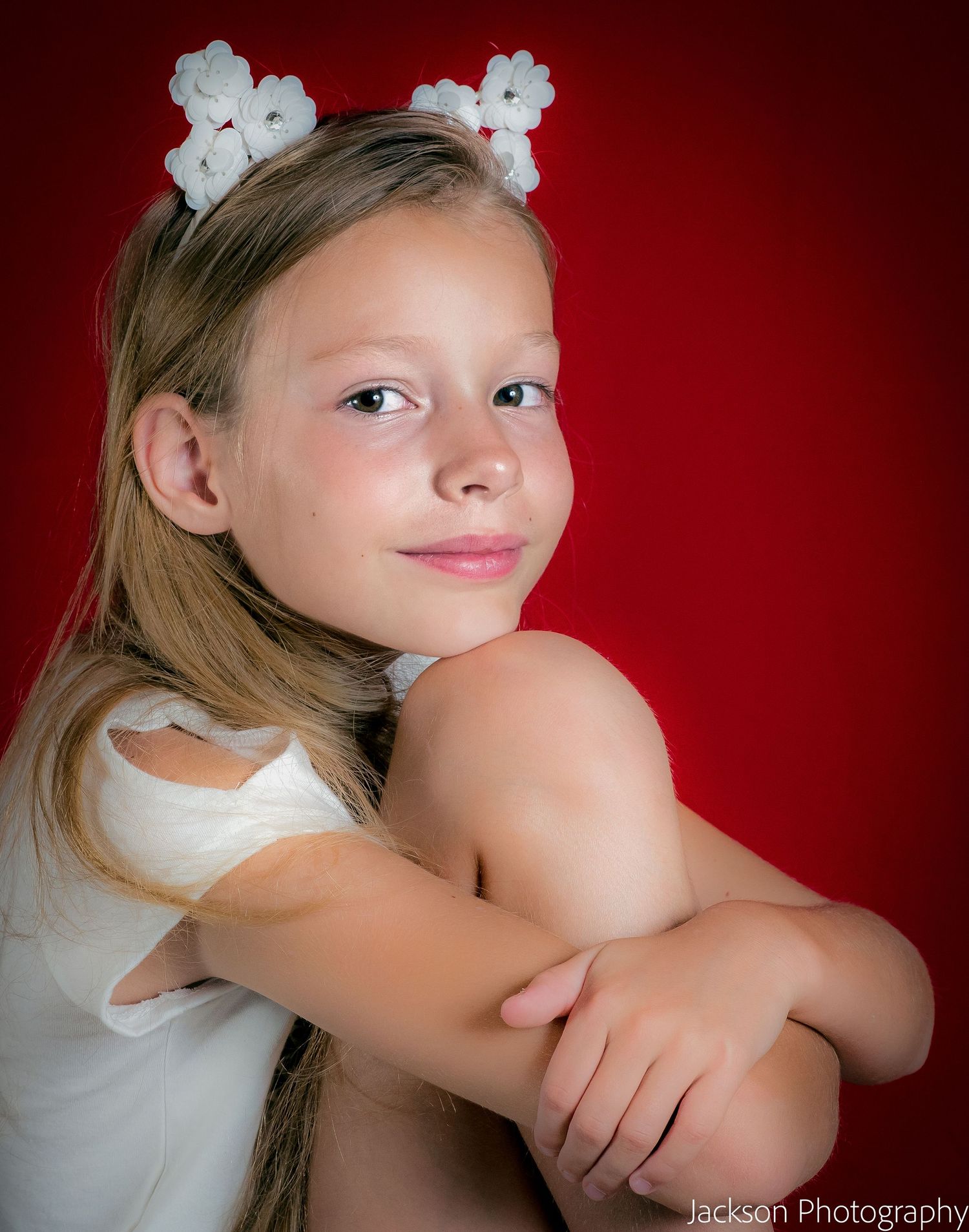 Young girl with white flower hair accessories poses gracefully against a vibrant red background in a professional portrait.