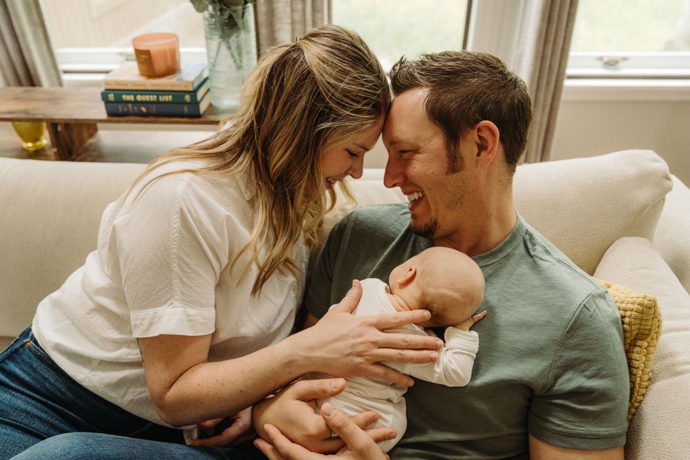 Parents sharing intimate moment while holding their newborn on couch in living room.