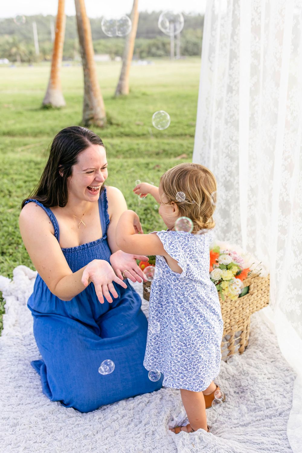 A laughing mother in blue dress plays with her toddler daughter amid bubbles on a white blanket outdoors.