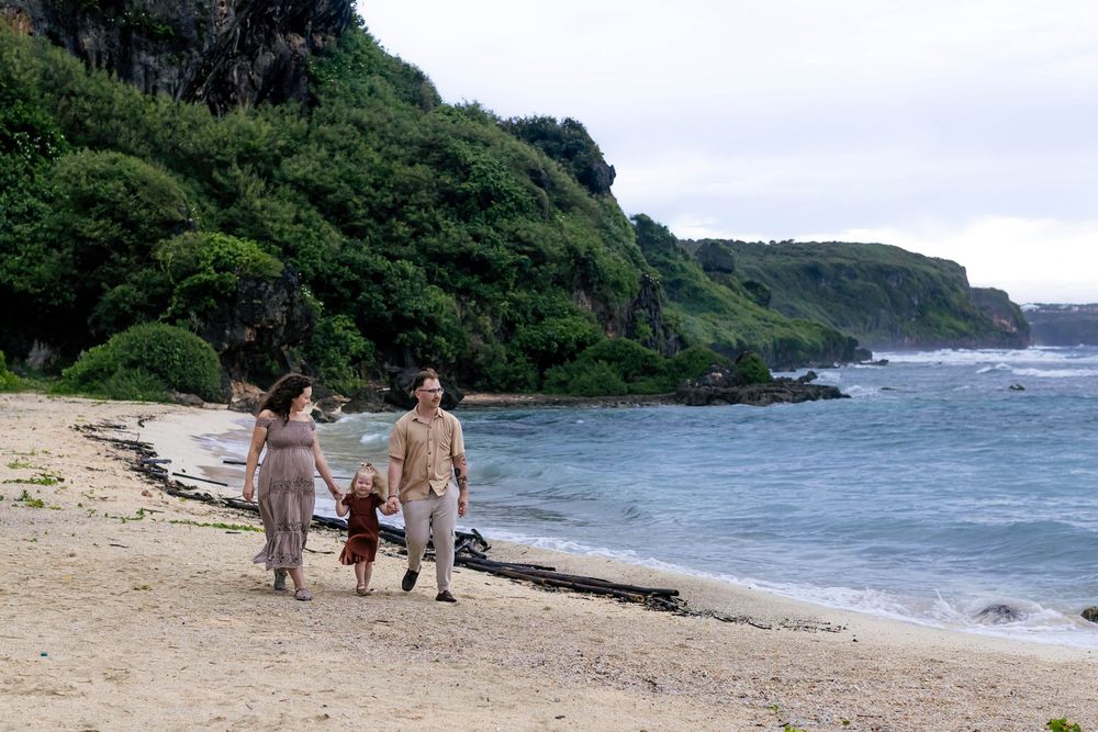 Three people walking on a sandy beach with green cliffs and ocean waves in the background.