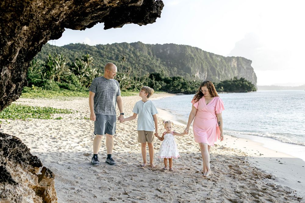 Family of four holding hands walking on a sandy tropical beach with lush green mountains in the background.