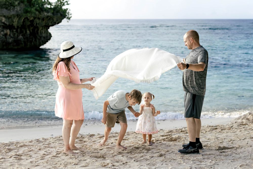 Family of four playing with white fabric on a sandy beach with turquoise ocean waves in the background.