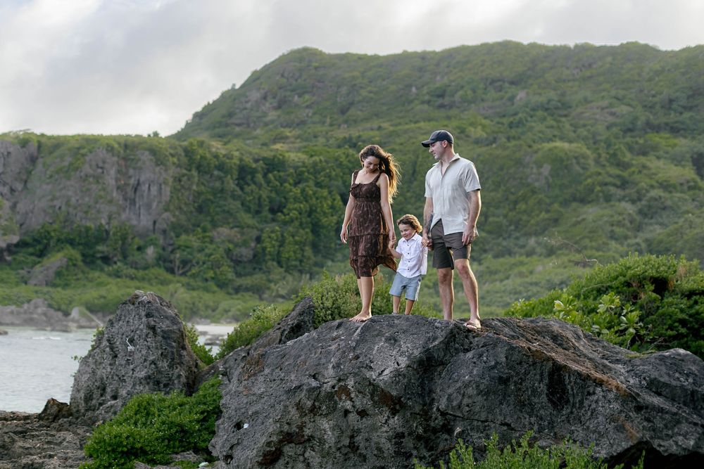 Family of three standing on rocky shore with lush green mountains and ocean in the background.