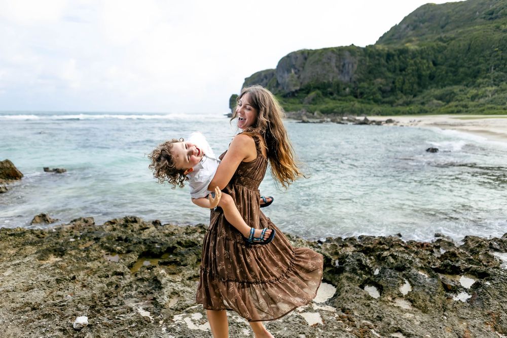 Woman in floral dress playfully lifting a child on a rocky beach with turquoise ocean and green mountains.