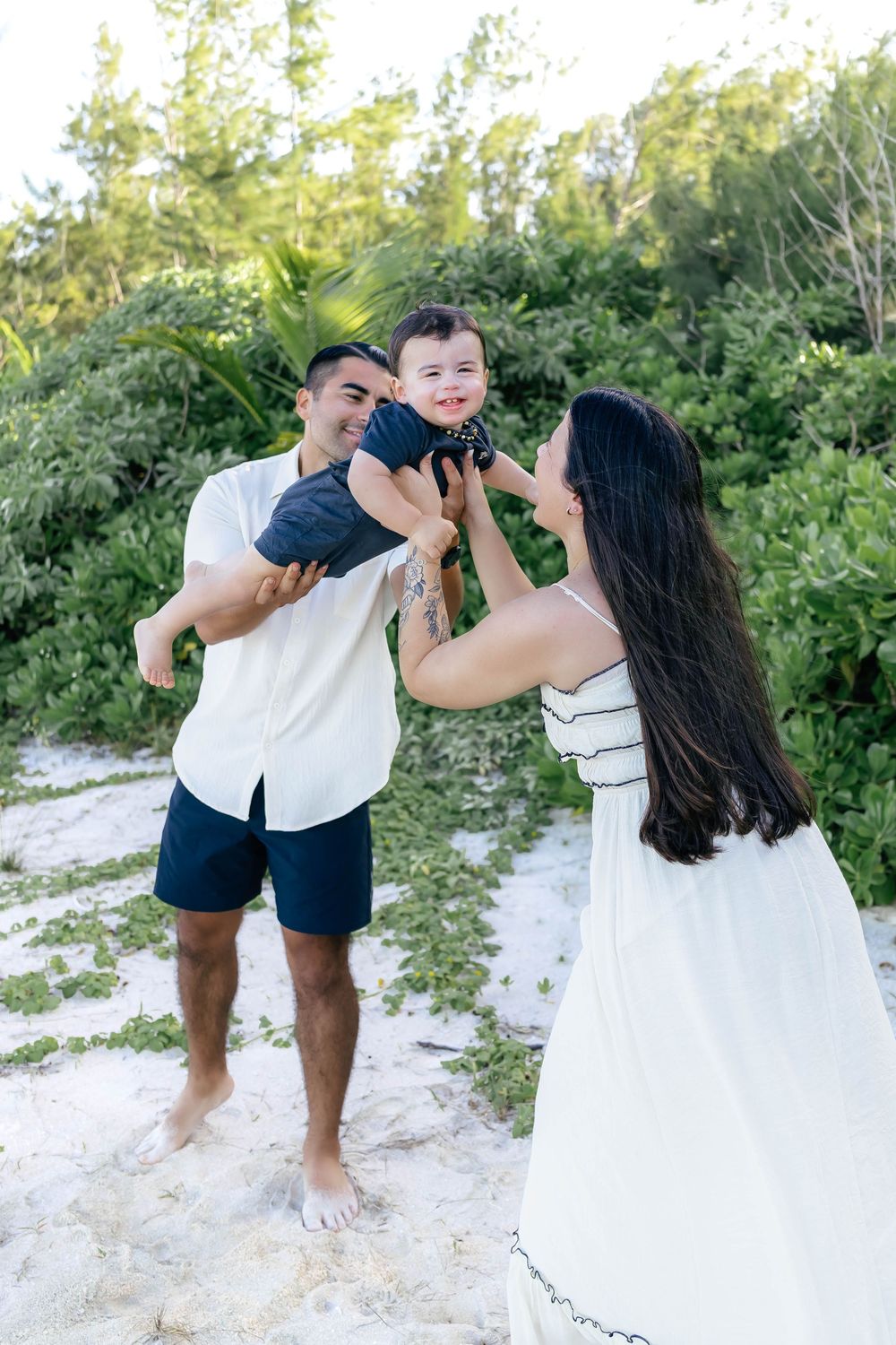 A happy family with a man and woman lifting their smiling baby outdoors on a sandy beach with lush greenery.