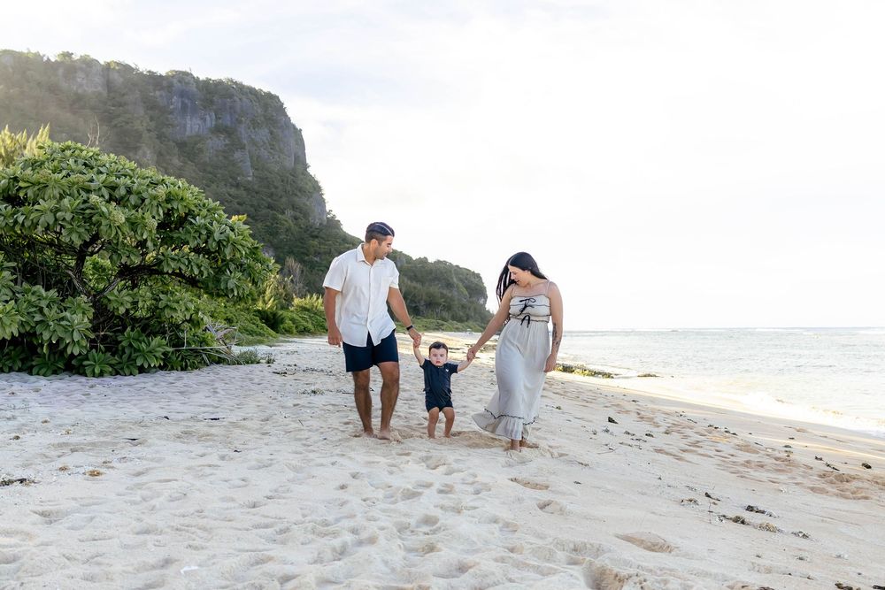 Family of three walking on a sandy beach with lush greenery and cliffs in the background.